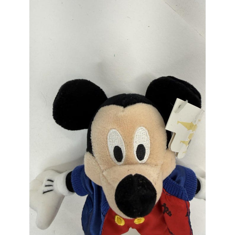 Peut inclure: Une peluche Mickey Mouse avec des oreilles noires, un visage beige et un nez noir. La poup&eacute;e porte une veste bleue, un pantalon rouge et des gants blancs. Une &eacute;tiquette est attach&eacute;e.
