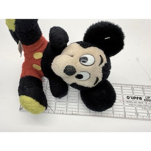 May include: A vintage Mickey Mouse plush toy, featuring a black body, red shorts with yellow buttons, and a cream-colored face. The toy is lying on a white surface next to a ruler.