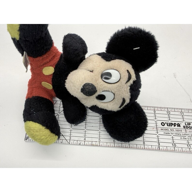 May include: A vintage Mickey Mouse plush toy, featuring a black body, red shorts with yellow buttons, and a cream-colored face. The toy is lying on a white surface next to a ruler.
