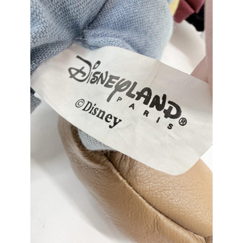 K&ouml;nnte beinhalten: Nahaufnahme eines DisneyLand Paris-Etiketts auf einem Pl&uuml;schtier. Das Etikett ist wei&szlig; mit schwarzem Text, der "DisneyLand Paris" und "&copy; Disney" lautet. Das Spielzeug hat Jeansstoff und einen hellbraunen Lederschuh.
