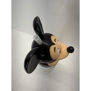 May include: A Mickey Mouse-shaped tumbler with a black head and ears, a tan face, and a black nose. The tumbler has a white straw and a white and black cup. The cup has a design of Mickey Mouse.