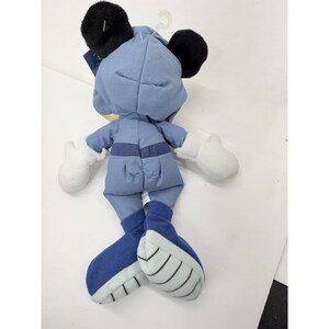 May include: A plush toy of a character wearing a light blue outfit with black ears and white gloves. The character's feet are dark blue with white accents. The toy is hanging from a white plastic hook.