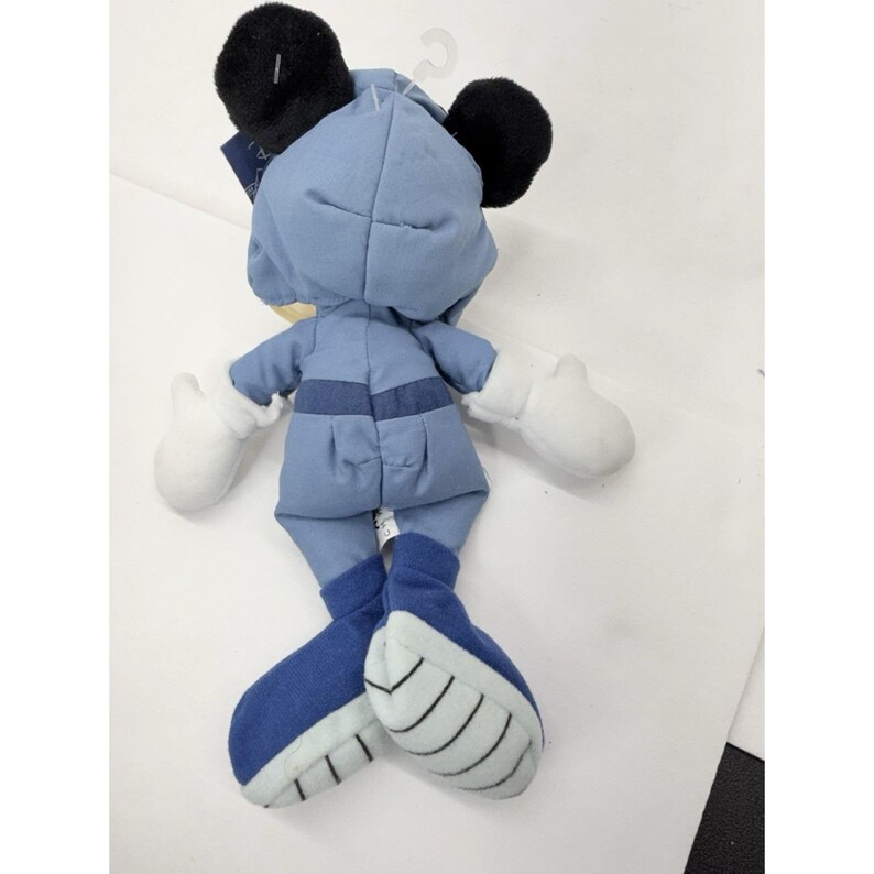 May include: A plush toy of a character wearing a light blue outfit with black ears and white gloves. The character's feet are dark blue with white accents. The toy is hanging from a white plastic hook.
