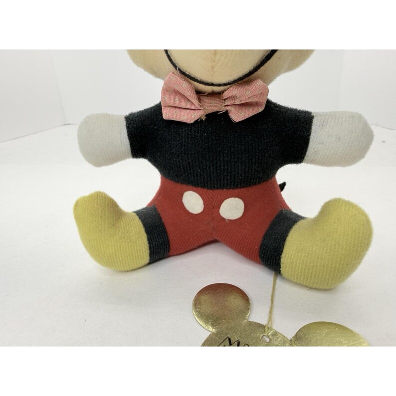 Puede incluir: Un peluche de Mickey Mouse con torso negro, pantalones cortos rojos y pies amarillos. El mu&ntilde;eco tiene una pajarita rosa y guantes blancos. Una etiqueta dorada est&aacute; unida al mu&ntilde;eco.