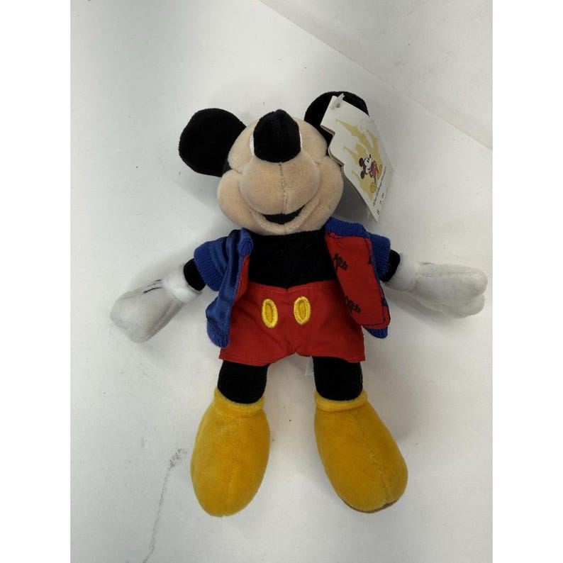 Peut inclure: Peluche de Mickey Mouse avec une veste bleue, un short rouge et des chaussures jaunes. La peluche a des oreilles noires, un nez noir et des gants blancs. Une &eacute;tiquette est attach&eacute;e.