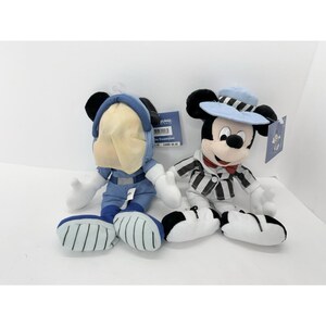 May include: Two plush Mickey Mouse dolls. One is dressed in a blue space suit, the other in a striped jacket and a light blue hat. Both have tags attached.