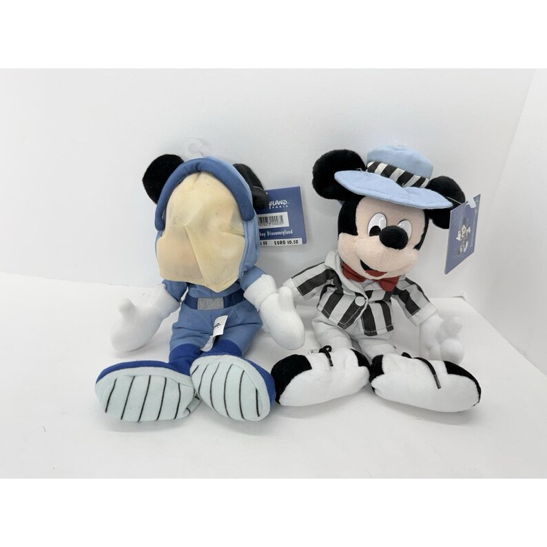 May include: Two plush Mickey Mouse dolls. One is dressed in a blue space suit, the other in a striped jacket and a light blue hat. Both have tags attached.