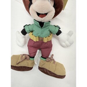 K&ouml;nnte beinhalten: Ein Pl&uuml;schtier von Mickey Mouse mit einem braunen Hut, einem gr&uuml;nen Hemd, einer weinroten Hose und beigefarbenen Schuhen. Das Spielzeug hat wei&szlig;e Handschuhe und einen gelben G&uuml;rtel. Das Mickey Mouse-Spielzeug ist ein Sammlerst&uuml;ck.