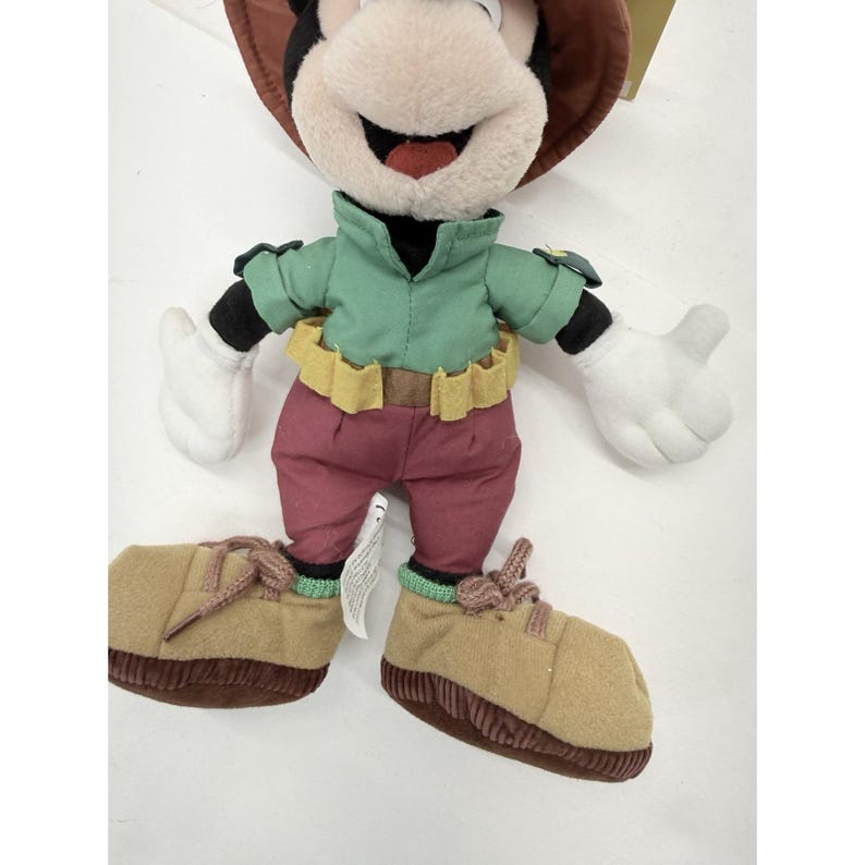 K&ouml;nnte beinhalten: Ein Pl&uuml;schtier von Mickey Mouse mit einem braunen Hut, einem gr&uuml;nen Hemd, einer weinroten Hose und beigefarbenen Schuhen. Das Spielzeug hat wei&szlig;e Handschuhe und einen gelben G&uuml;rtel. Das Mickey Mouse-Spielzeug ist ein Sammlerst&uuml;ck.