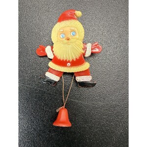 May include: A vintage-style Santa Claus ornament featuring a red suit with cream-colored trim, a white beard, blue eyes, and black boots. A small red bell hangs from the bottom of the figure by a thin string, creating a festive hanging decoration.