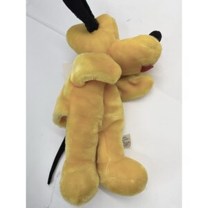 Puede incluir: Un peluche amarillo de Pluto, el perro, con orejas y cola negras. El juguete tiene una lengua roja y una nariz negra. Est&aacute; hecho de un material suave y difuso y tiene una etiqueta en una pata. Un objeto de colecci&oacute;n vintage de Disney.