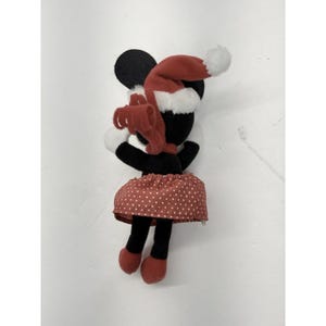 May include: A Minnie Mouse plush toy wearing a red Santa hat with a white pom-pom and a red polka dot skirt. The plush has black ears, a black head, and black legs with red shoes. A red bow is around the neck.