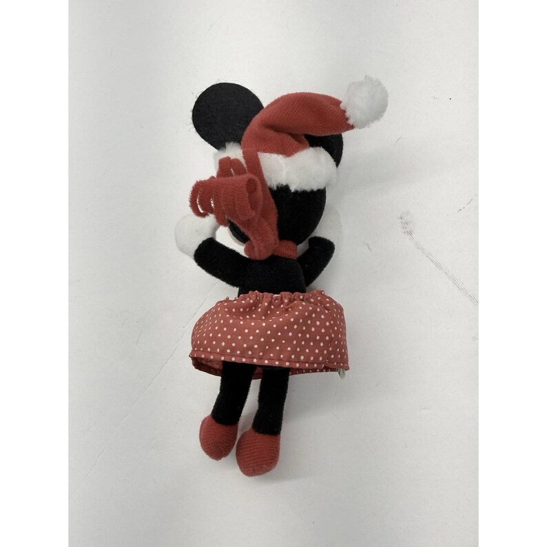May include: A Minnie Mouse plush toy wearing a red Santa hat with a white pom-pom and a red polka dot skirt. The plush has black ears, a black head, and black legs with red shoes. A red bow is around the neck.