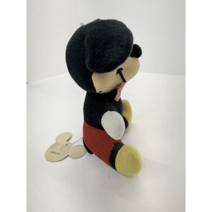Puede incluir: Un peluche vintage de Mickey Mouse. El peluche presenta una cabeza y orejas negras, un cuerpo negro, pantalones cortos rojos, pies amarillos y una pajarita rosa. Una etiqueta est&aacute; adjunta. El juguete est&aacute; en posici&oacute;n sentada.