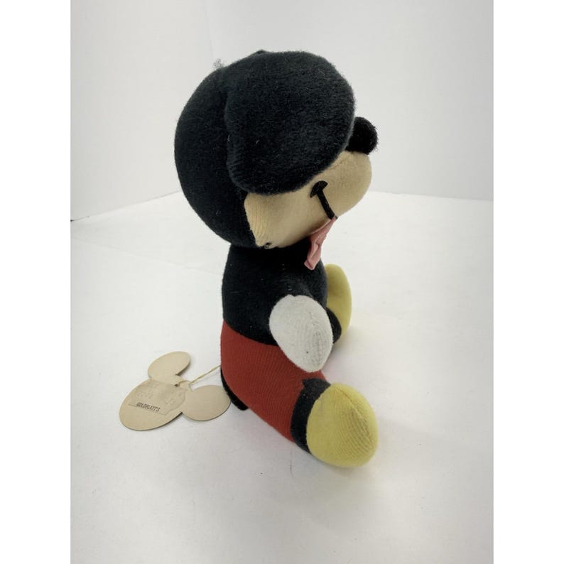 Puede incluir: Un peluche vintage de Mickey Mouse. El peluche presenta una cabeza y orejas negras, un cuerpo negro, pantalones cortos rojos, pies amarillos y una pajarita rosa. Una etiqueta est&aacute; adjunta. El juguete est&aacute; en posici&oacute;n sentada.