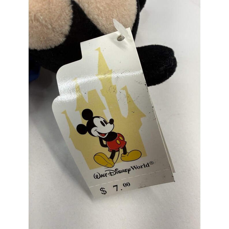 Peut inclure: Une peluche Mickey Mouse avec une &eacute;tiquette repr&eacute;sentant le logo Walt Disney World et une image de dessin anim&eacute; de Mickey Mouse. L'&eacute;tiquette a une silhouette de ch&acirc;teau en arri&egrave;re-plan. Le jouet est noir, beige, rouge et jaune.