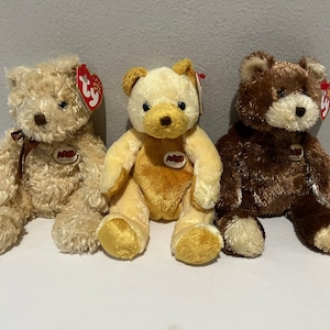 May include: Three plush teddy bears in varying shades of brown and tan. Each bear has a Ty tag, a brown nose, and a small ribbon or patch. The bears are sitting side by side.