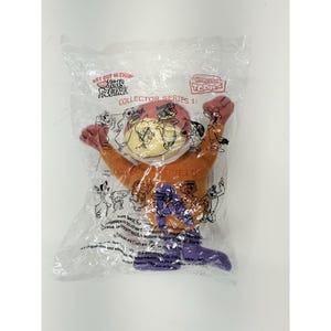 May include: A plush toy of a cartoon character in a clear plastic bag. The toy is orange, purple, and pink, with a round head and outstretched arms. The packaging includes text that reads "Collector Series 1:" and "Kids Meal".