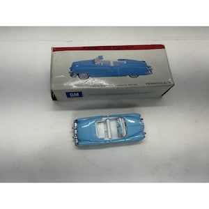May include: A light blue 1953 Cadillac Eldorado toy car with a white interior, displayed next to its original box. The box features an image of the car and the text "Cadillac 1953 Eldorado". The car is a miniature replica.