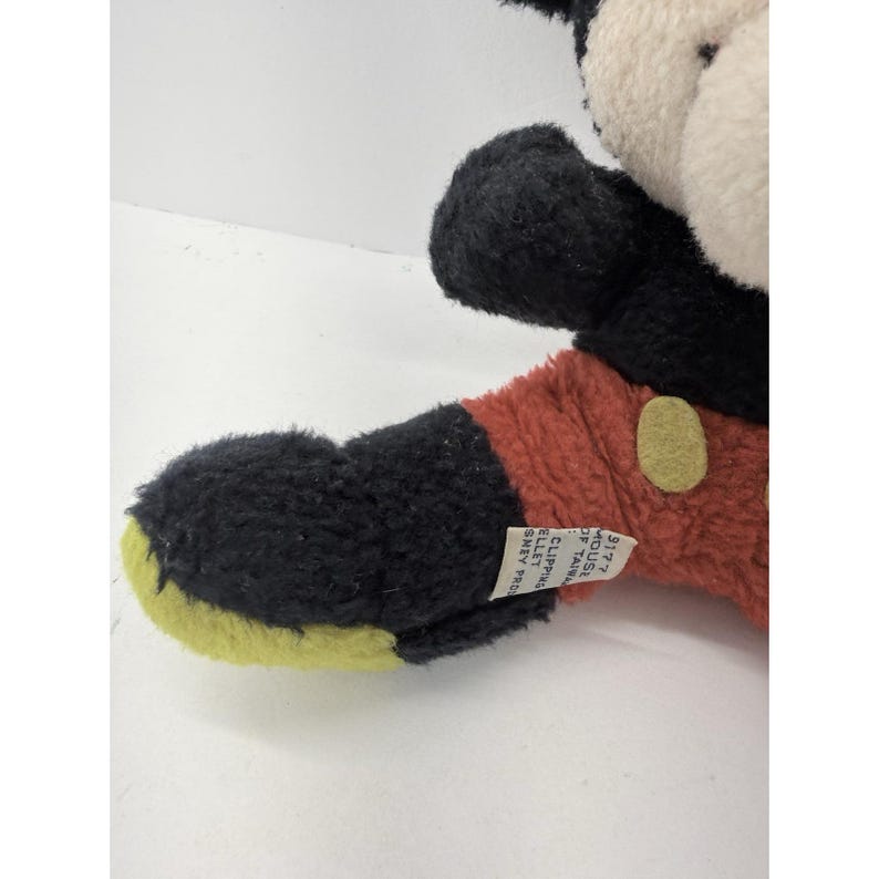 May include: A vintage Mickey Mouse plush toy. The plush features black ears, a black head, and a white face. The body is red with black arms and yellow feet. A small tag is attached to the arm.