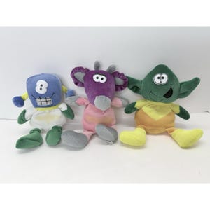 May include: Three plush toys in a row. The first is blue with a white belly, green feet, and a smiling mouth. The second is purple with a pink body and gray feet. The third is green with yellow accents.