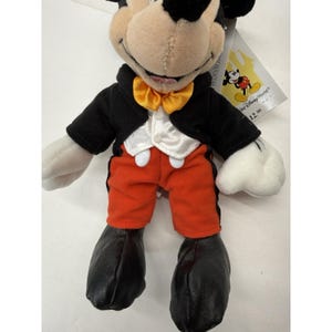 Peut inclure: Peluche de Mickey Mouse portant une veste de smoking noire, un short orange et des chaussures noires. Le jouet a un n&oelig;ud papillon jaune et des gants blancs. Une &eacute;tiquette Disney World est attach&eacute;e.