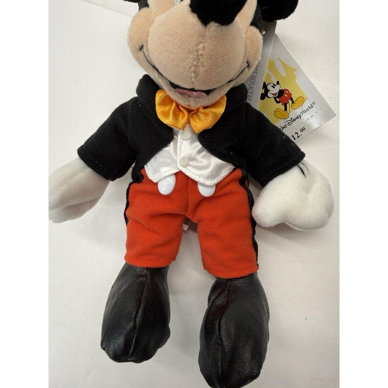 Peut inclure: Peluche de Mickey Mouse portant une veste de smoking noire, un short orange et des chaussures noires. Le jouet a un n&oelig;ud papillon jaune et des gants blancs. Une &eacute;tiquette Disney World est attach&eacute;e.