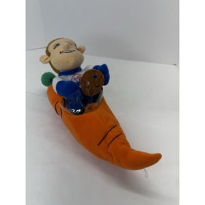 May include: A plush toy featuring a cartoon monkey wearing a blue and white racing suit, seated in an orange carrot-shaped vehicle. The monkey has a smiling expression. The carrot vehicle has a clear plastic windshield.