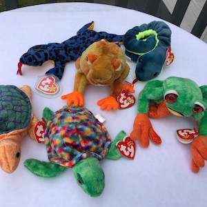 May include: Five colorful Beanie Baby plush toys: a green and purple turtle, a green and orange turtle, a blue and black lizard, a green frog with orange feet, and a blue snake with a yellow stripe.