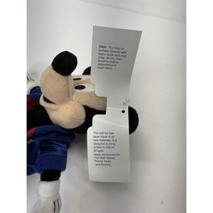 Peut inclure: Peluche Mickey Mouse avec un visage cr&egrave;me, des oreilles noires et une veste bleue. Le jouet a une &eacute;tiquette avec des instructions d'entretien et le texte : "Ce jouet en peluche a &eacute;t&eacute; fabriqu&eacute; avec de nouveaux mat&eacute;riaux."