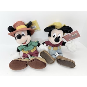 K&ouml;nnte beinhalten: Zwei Mickey Mouse-Pl&uuml;schtiere in Cowboy-Outfits. Einer tr&auml;gt ein gr&uuml;nes Hemd und eine weinrote Hose, der andere ein rotes Hemd und blaue Jeans. Beide haben braune Schuhe und wei&szlig;e Handschuhe.