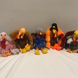 May include: A collection of five plush turkey toys in various colors. The turkeys have different colored feathers, beaks, and feet. One turkey wears a pilgrim hat. The toys are arranged on a white surface.