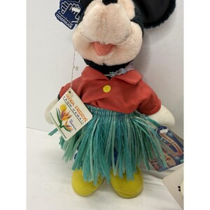 Puede incluir: Un mu&ntilde;eco de peluche de Mickey Mouse con una camisa roja, una falda de hierba verde y zapatos amarillos. El mu&ntilde;eco tiene orejas negras y una cara blanca. Una etiqueta dice "Aloha Friends From Hawaii!" y "by applause".