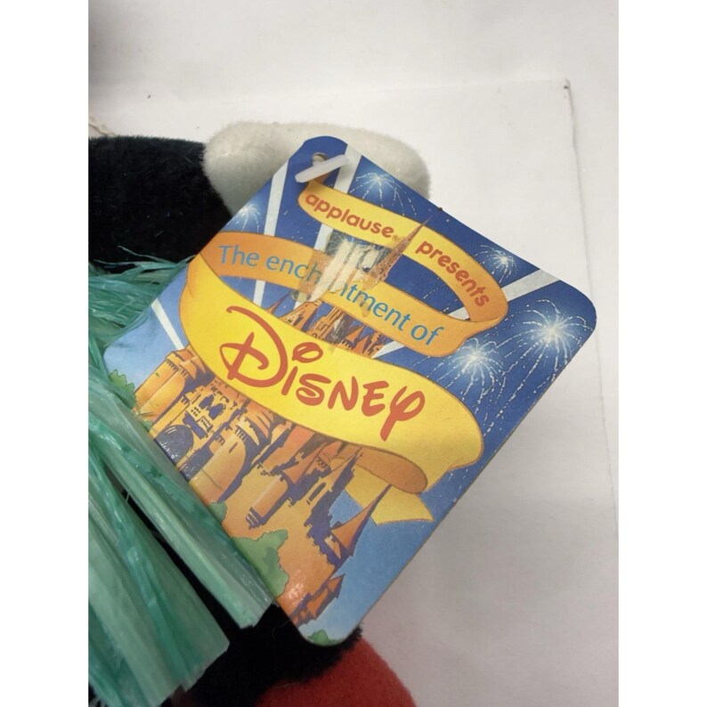 Puede incluir: Una etiqueta de producto de Disney con las palabras "applause presents" y "The enchantment of Disney" impresas. La etiqueta tiene un fondo azul con fuegos artificiales y una ilustraci&oacute;n de castillo. La etiqueta est&aacute; adjunta a un peluche.