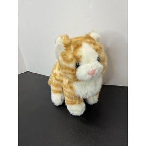 May include: A plush cat toy featuring orange and white striped fur. The stuffed animal has a white chest, white paws, and a white face with a small pink nose. It has dark eyes and pointed ears, with a soft, fuzzy texture throughout its body.