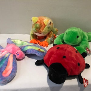 May include: Four colorful plush toys: a green frog, a red ladybug, a purple and green butterfly, and an orange and green frog. The toys are all lying on a white surface.