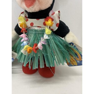 Puede incluir: Un peluche de un personaje en blanco y negro que lleva un vestido blanco con lunares rojos, una falda de hierba verde y un collar de flores de colores. El personaje tiene zapatos rojos y sostiene una etiqueta.
