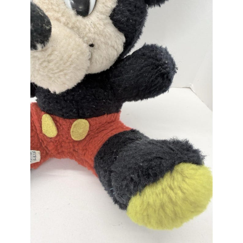 May include: Vintage Mickey Mouse plush toy. The plush features a black body, red shorts with yellow buttons, and yellow feet. The toy's face is white with black eyes and a black nose. A tag is attached to the shorts.