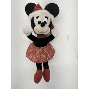 May include: A Minnie Mouse plush toy wearing a red and white Santa hat, a red scarf, and a red polka dot skirt. The plush has black ears, a black body, white gloves, and red shoes. The toy is designed for the holiday season.