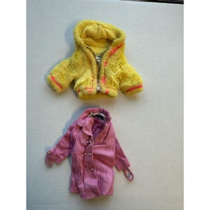 May include: Two doll-sized jackets. The top jacket is yellow with a hood, a silver zipper, and pink trim. The bottom jacket is pink with silver buttons and a collar. Both jackets are on a white surface.