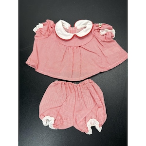 May include: A red and white gingham doll outfit consisting of a short-sleeved blouse and matching bloomers. The blouse features a white Peter Pan collar with red trim and white lace sleeve cuffs. The bloomers have white lace trim at the leg openings.