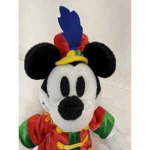 Puede incluir: Un peluche de Mickey Mouse con un atuendo rojo y verde, un sombrero amarillo y una pluma azul. El juguete tiene orejas negras y una cara blanca. Un objeto de colecci&oacute;n de Disney.