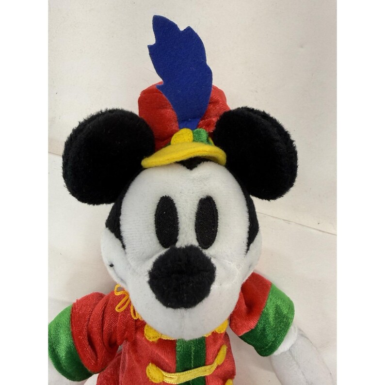 Puede incluir: Un peluche de Mickey Mouse con un atuendo rojo y verde, un sombrero amarillo y una pluma azul. El juguete tiene orejas negras y una cara blanca. Un objeto de colecci&oacute;n de Disney.