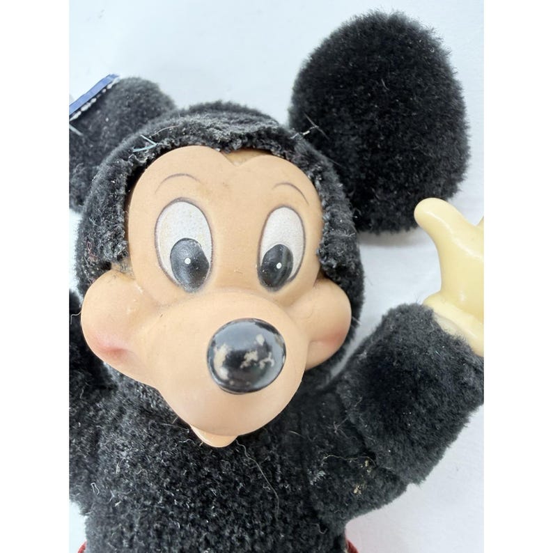 May include: Vintage Mickey Mouse plush toy with black fur, a tan face, and large, expressive eyes. The toy has a black nose and a red bottom. One hand is raised in a friendly gesture. A classic Disney collectible.