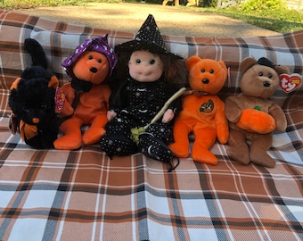 May include: A collection of Halloween-themed plush toys, including a black cat, orange bears, and a witch doll. The bears wear witch hats and carry pumpkins. The toys are arranged on a brown and white plaid blanket.