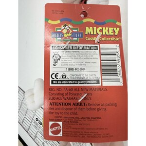Puede incluir: Una etiqueta roja de Mickey Mouse "Cuddy Collectible" con informaci&oacute;n para el consumidor, incluido un n&uacute;mero de tel&eacute;fono y certificaciones de seguridad. La etiqueta est&aacute; adherida a un juguete de peluche blanco. El texto "Mickey's Stuff for Kids" es visible.
