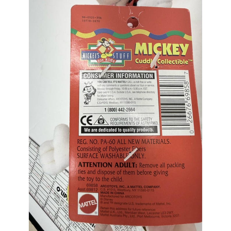 Puede incluir: Una etiqueta roja de Mickey Mouse "Cuddy Collectible" con informaci&oacute;n para el consumidor, incluido un n&uacute;mero de tel&eacute;fono y certificaciones de seguridad. La etiqueta est&aacute; adherida a un juguete de peluche blanco. El texto "Mickey's Stuff for Kids" es visible.