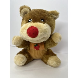 May include: A brown plush teddy bear with a red strawberry nose and a yellow corduroy bib with a strawberry design.