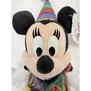 May include: A plush Minnie Mouse doll wearing a party hat with red, green, purple, and yellow accents. The doll has black ears, large white eyes, and a pink face. She is wearing a green, purple, and red outfit.