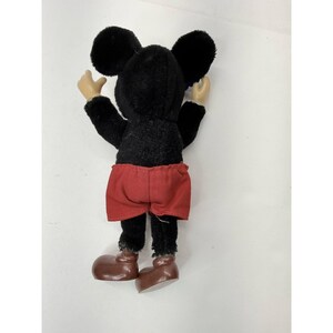 May include: A vintage Mickey Mouse plush toy, viewed from the back. The plush features a black body, large round ears, red shorts, and brown shoes. The arms and hands are a light tan color. The toy is standing with its arms raised.